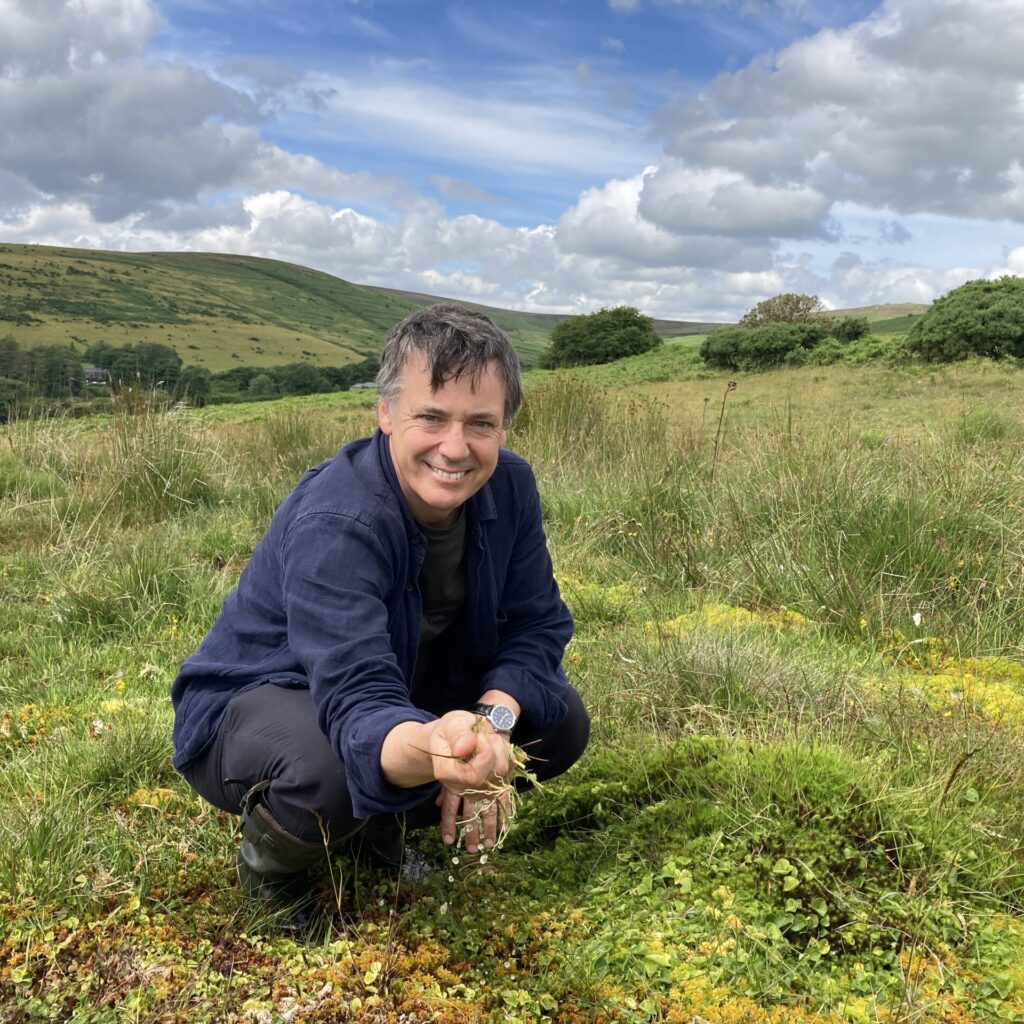 A John Wedgwood Clarke crouching with sphagnum moss in his hand, smiling into the camera.