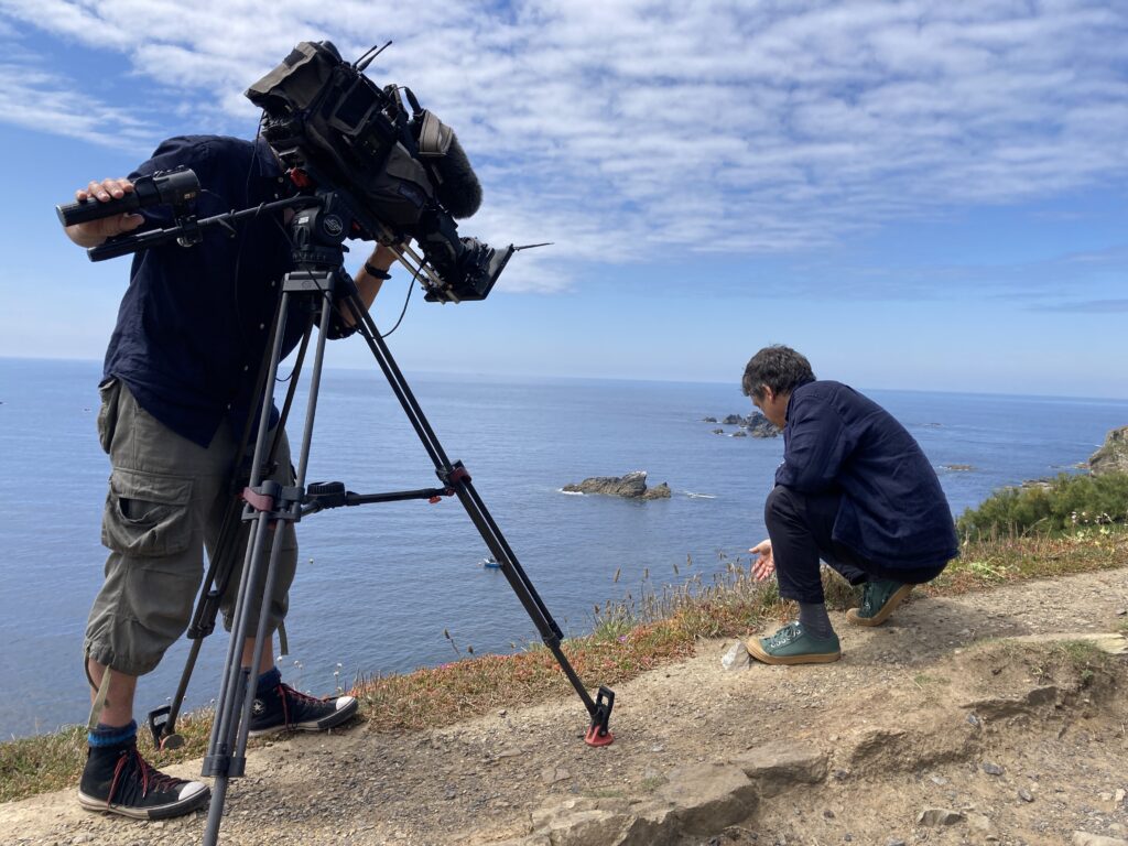 A man crouching on a coastal path, being filmed by another man with a camera