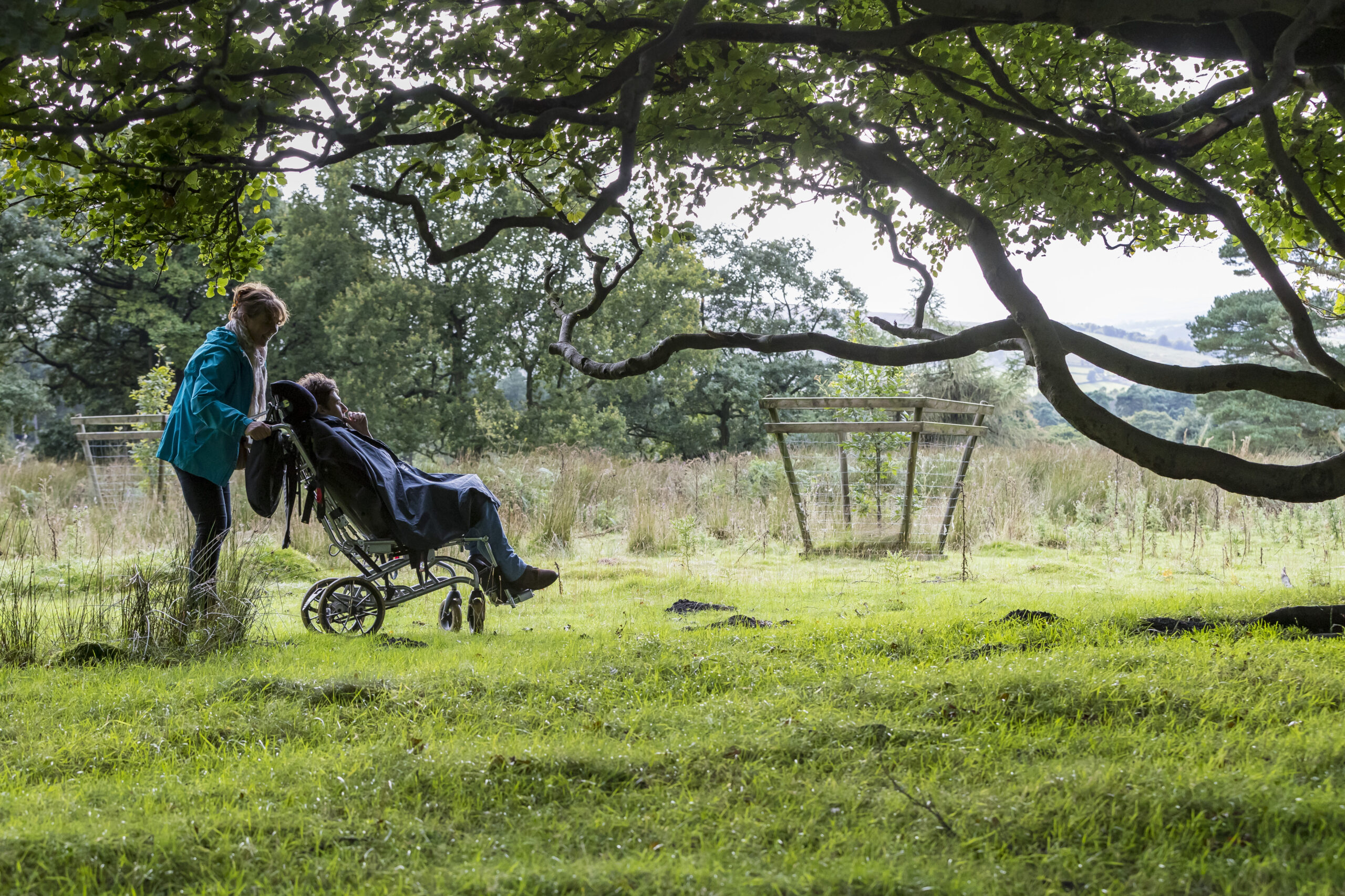A woman pushing a man in a wheelchair through a countryside landscape