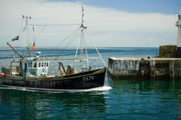 A fishing trawler entering a harbour, with lighthouse in the background