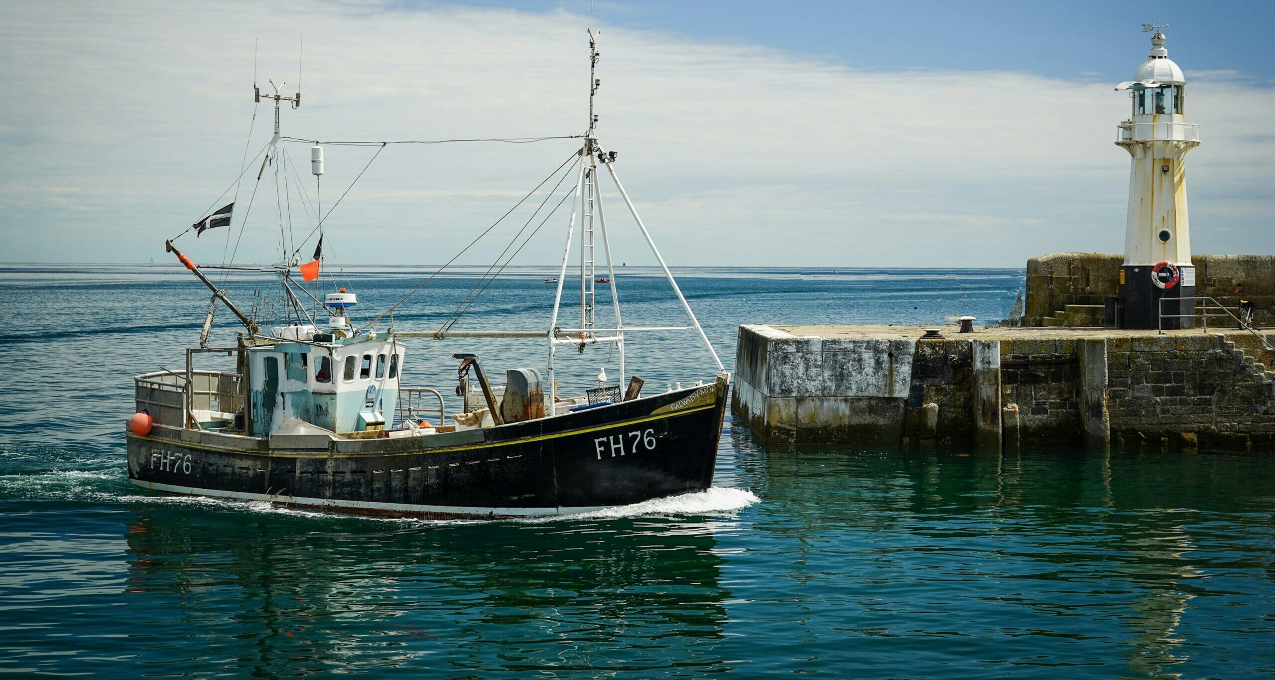 A fishing trawler entering a harbour, with lighthouse in the background